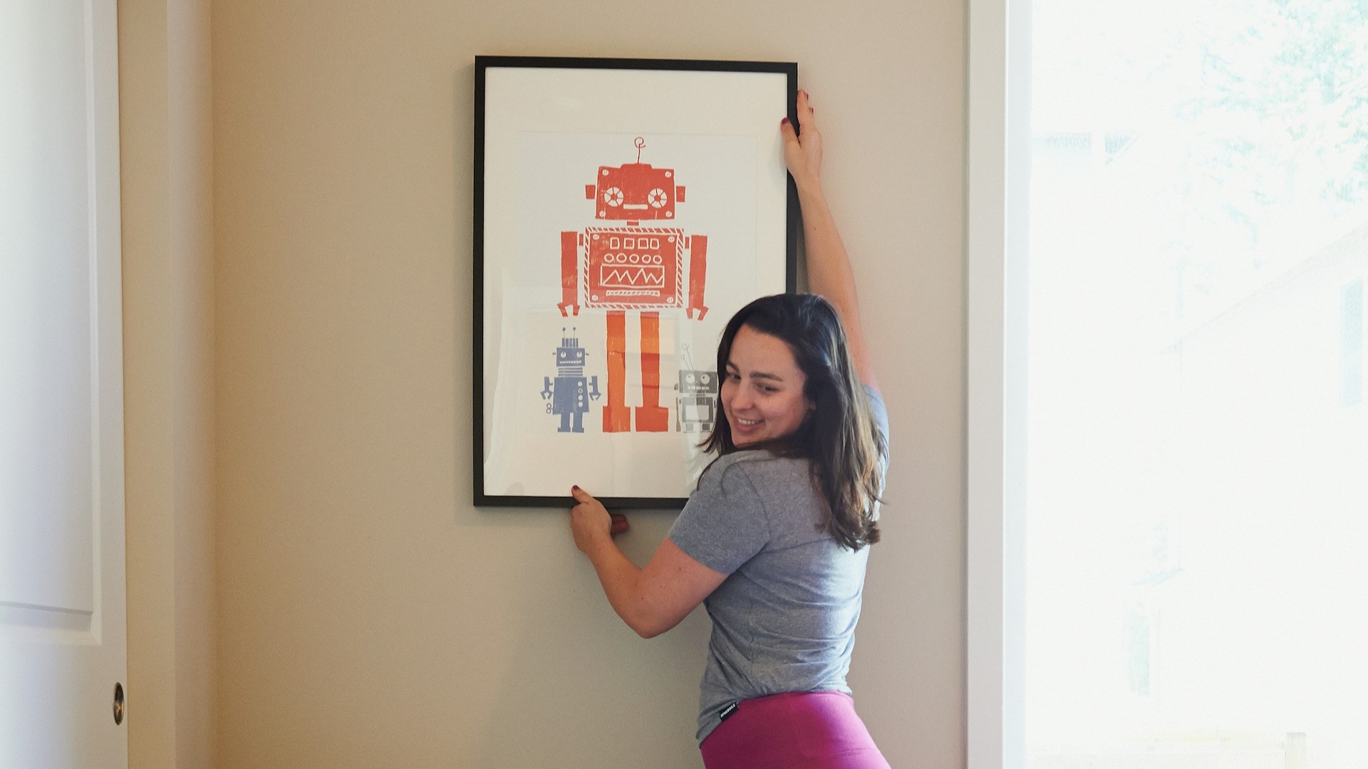 Vrouw hangt een ingelijst kunstwerk met drie kleurrijke robotillustraties op aan de muur