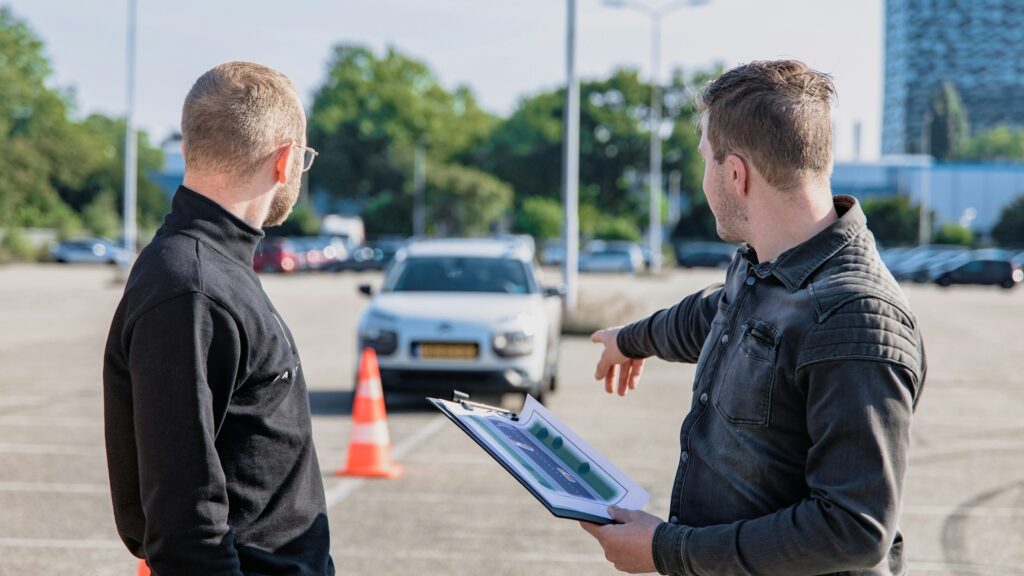 Rijinstructeur geeft uitleg naast leerling voor praktijkles met automaatrijbewijs op parkeerterrein.